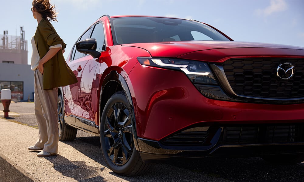 Close-up of the passenger-side front bumper of a parked Soul Red Crystal Metallic Mazda CX-5, with a woman standing nearby looking away.